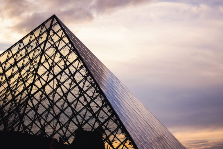 Vista exterior del Museo del Louvre al atardecer, con su icónica pirámide de cristal iluminada.